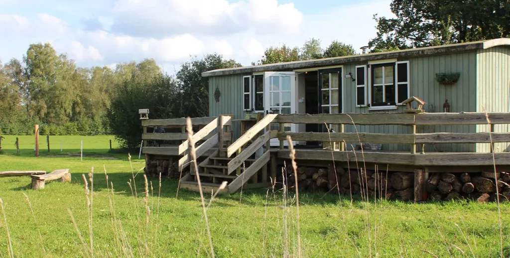 Lichtgroene houten pipowagen met een veranda, gelegen op een grasveld met omringende bomen. Er zijn houten trappen naar de veranda en stapels hout onder de wagen. Op de achtergrond is een weide te zien.